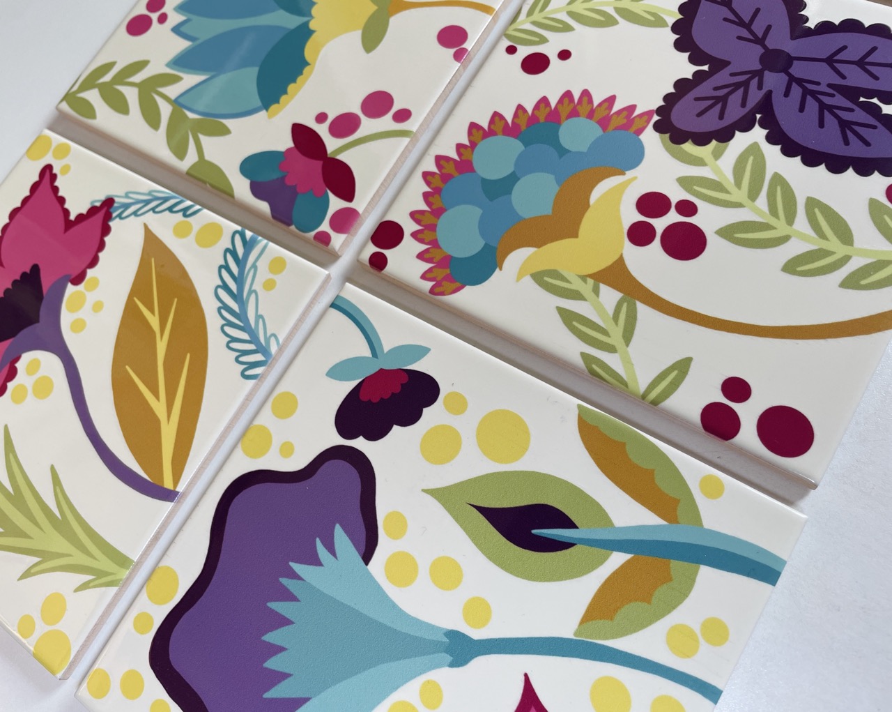 Four ceramic tiles arranged in a square, each featuring vibrant floral patterns in various colours, including pink, blue, green, and purple. The design incorporates leaves and berries against a cream background.