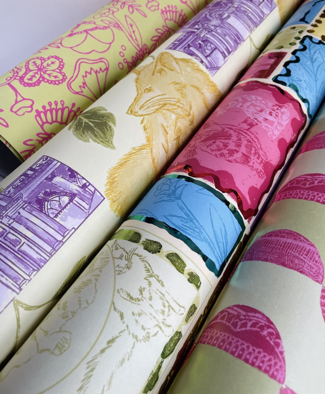 Five rolled sheets of patterned wrapping paper displayed together. The designs include colourful illustrations of foxes and cats, intricate floral motifs, and various geometric patterns in pink, blue, and yellow tones.