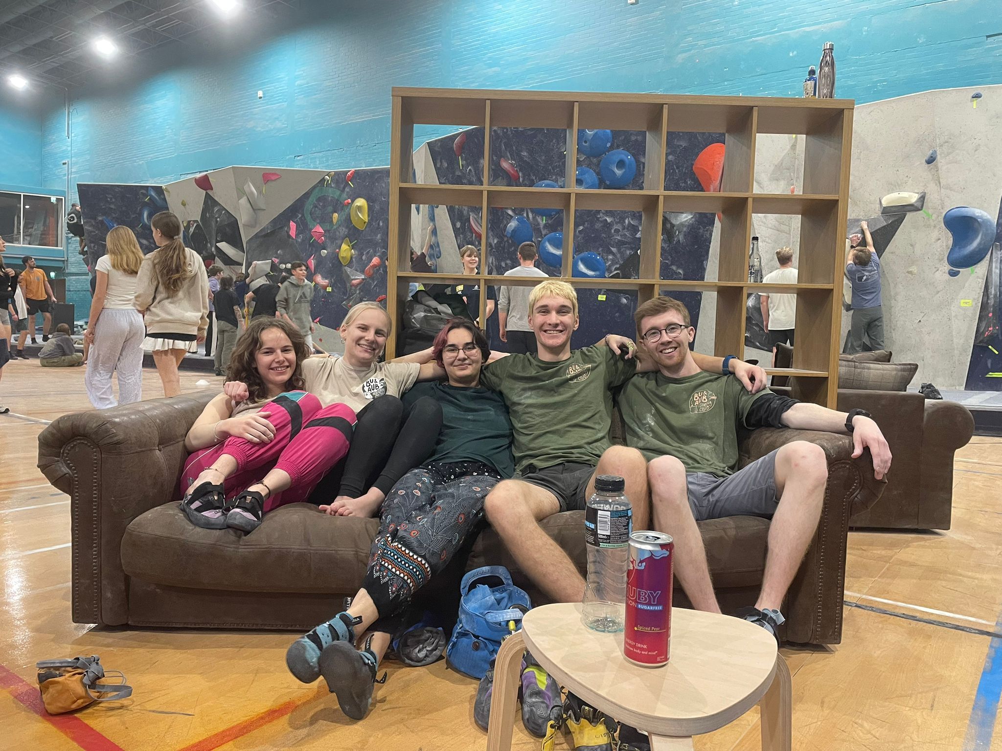 Five people sitting on brown sofa in climbing gym, storage cubbies with gear behind them, blue and grey climbing walls visible.