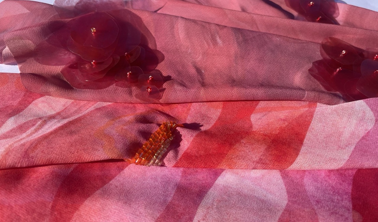A close-up view of layered fabric pieces in shades of pink and red. The top layer features textured red flowers, while the bottom layer displays a gradient pattern of pink tones with a subtle sheen.