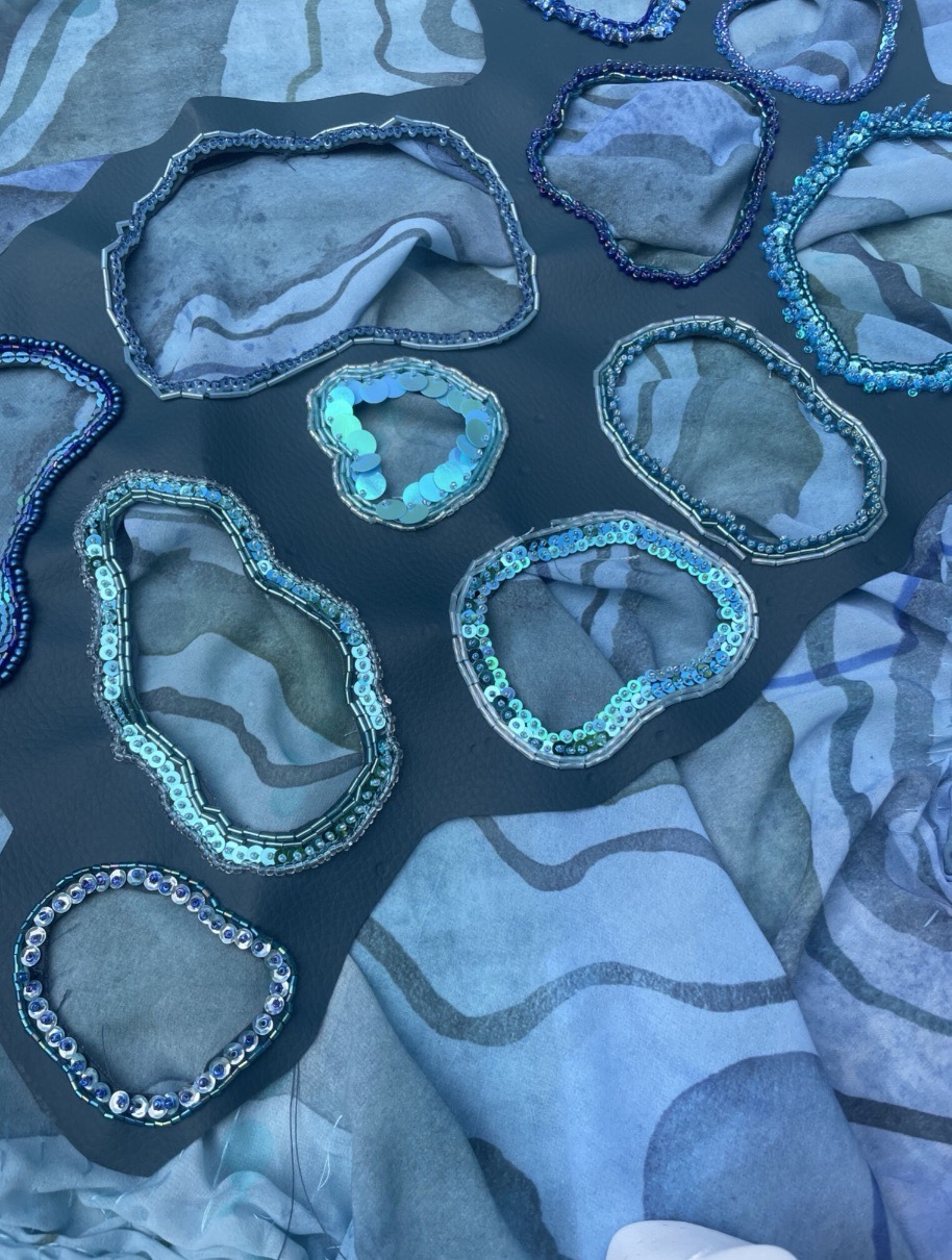 An assortment of blue sequins and fabric patches arranged on a textured blue and grey background. The sequins are outlined in wavy shapes, creating various sizes and depths against the underlying fabric.