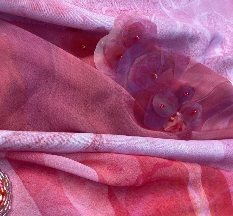 A close-up view of layered fabrics in shades of pink, featuring soft floral embellishments. The fabric has a smooth texture, and the flowers include small beads, creating a delicate and intricate design.
