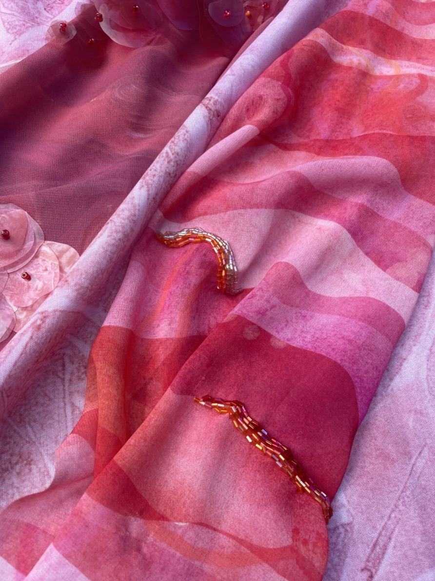 A close-up of layered fabrics in various shades of pink and red. Intricate embroidery in gold thread is visible on one fabric, while the background features smooth silk-like textures against a softly patterned textile.