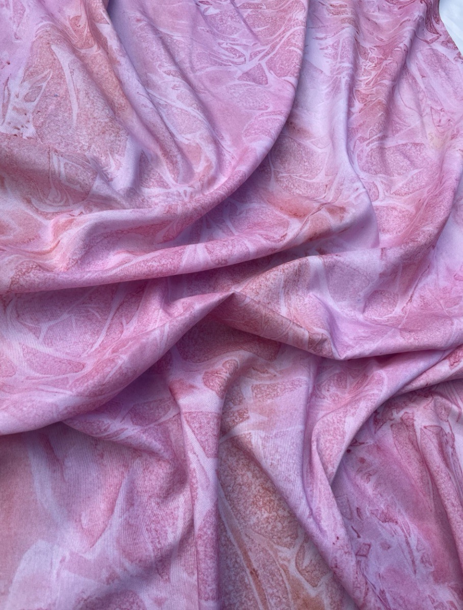 A draped piece of fabric in shades of pink, featuring a soft, textured pattern. The fabric appears to have a lightweight, flowing quality, with subtle variations in colour and design throughout.