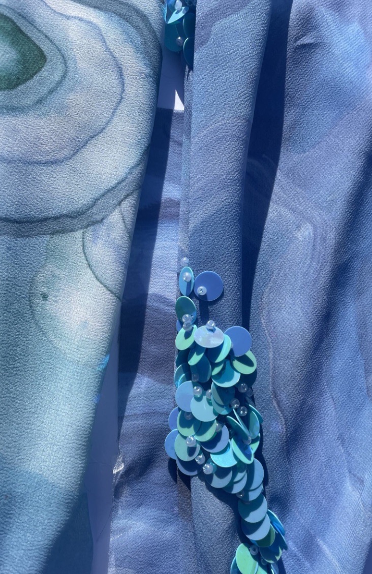 A close-up view of layered blue fabrics, featuring swirling patterns and textures. One side is adorned with shiny blue sequins, creating a contrast with the soft fabric surface.