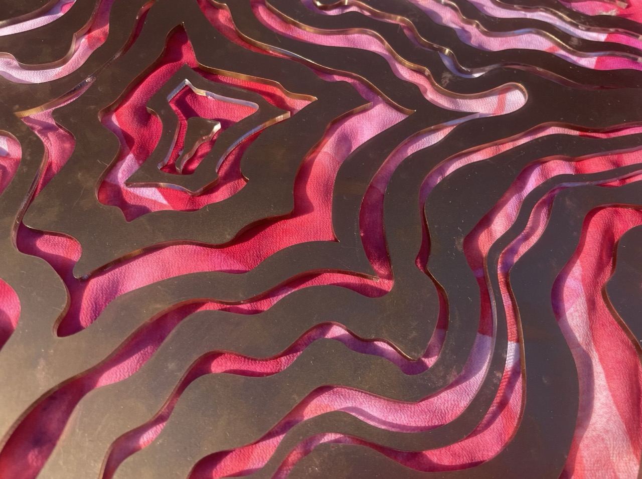 An abstract metal design with flowing, wavy patterns in various shapes. The metal surface is reflecting a pink fabric underneath, creating a layered effect with depth and texture.