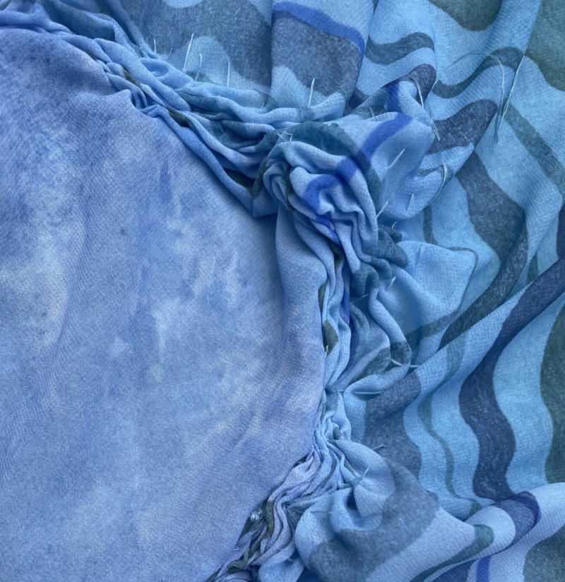 A close-up view of blue fabric featuring various shades and patterns, including wavy lines and soft textures. The fabric is gathered at the edges, showcasing its pliability and design.