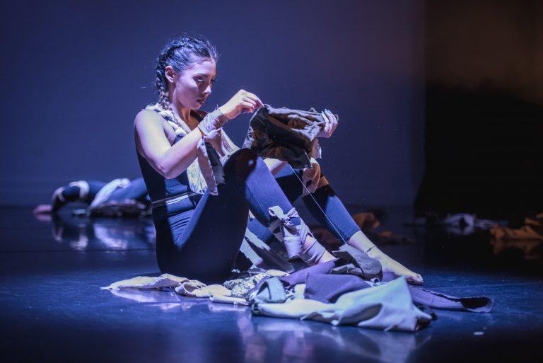 A dancer sits on stage feeling an object in her hands