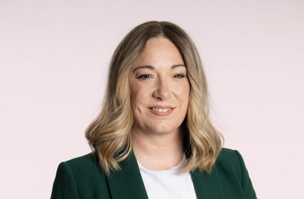 Woman with blonde wavy hair wearing green blazer over white top, smiling at camera against pale pink background.