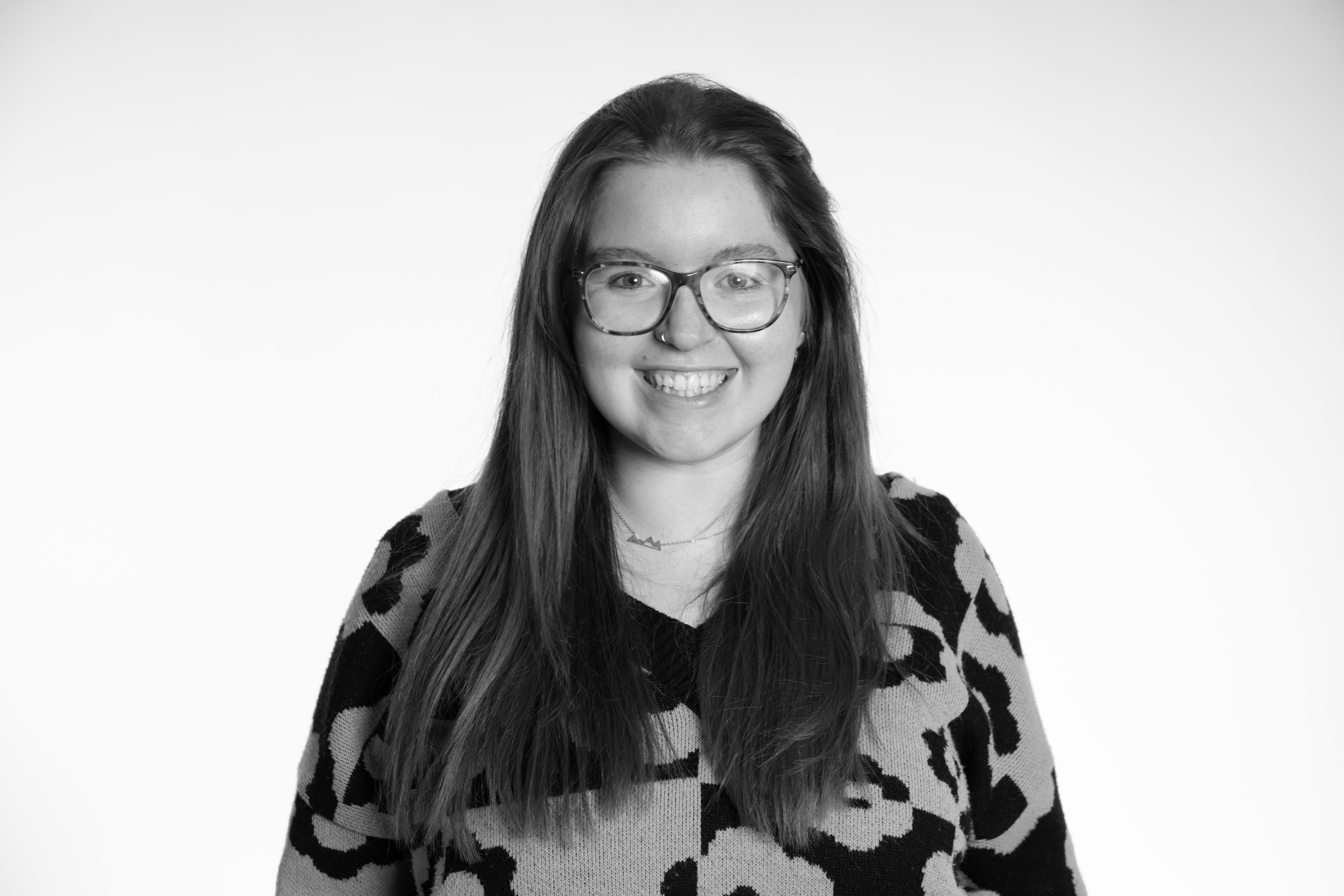 Black and white portrait of smiling woman with long hair, round glasses, wearing patterned top against plain background.