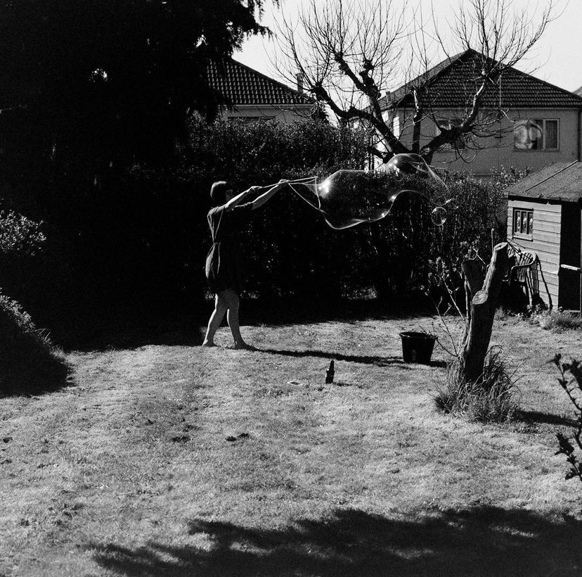 Black and white photo of a person making a large bubble in a back garden.