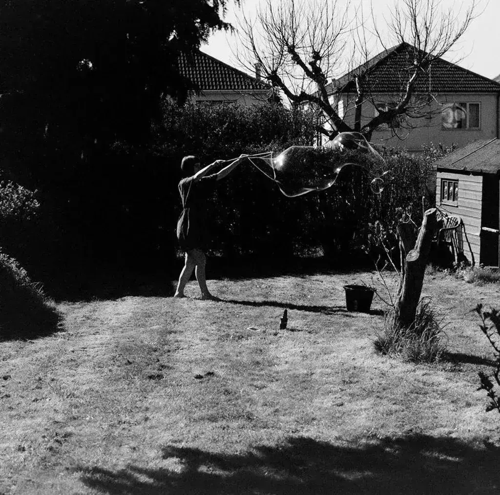 Black and white image showing person hanging washing on line in backyard with bare tree, houses with tiled roofs in background