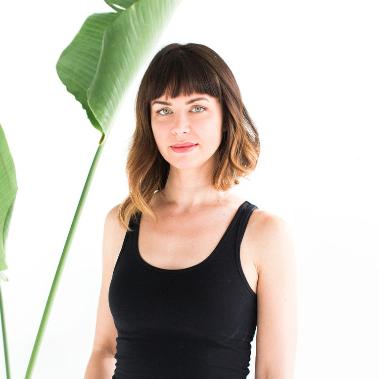 Woman with brown bob haircut wearing black sleeveless top against white background with large green tropical plant leaf visible on left side.