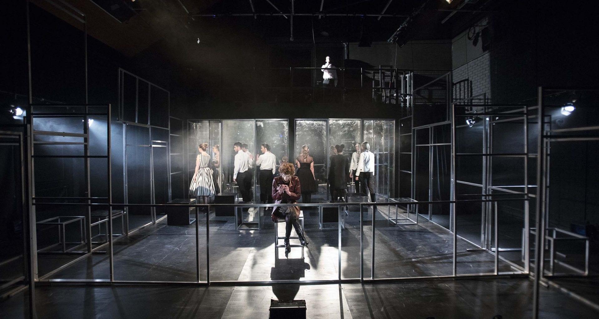 A person in costume sits cross-legged on a stool in the centre of a stage. Other people are behind them and facing towards a series of glass panels.