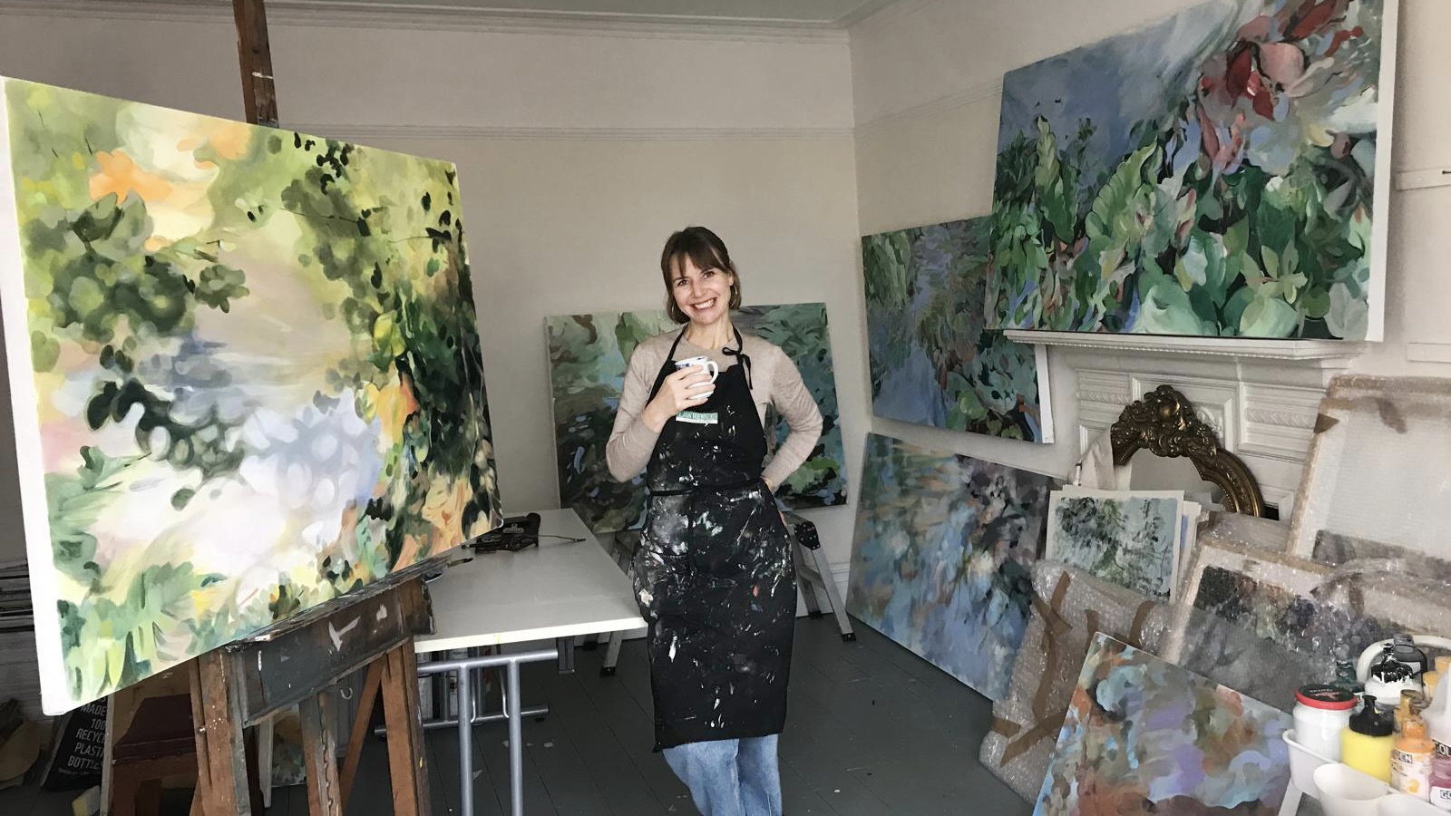 A woman in a paint-splattered apron stands in an art studio, surrounded by large floral and abstract canvases on easels and leaning against the walls.