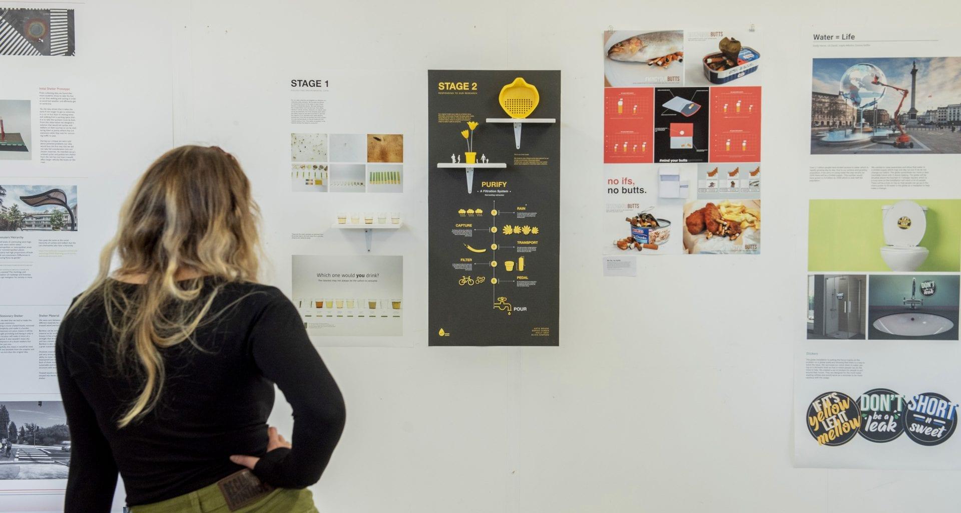 Pieces of graphic design work hang on a gallery wall. A person stands and looks at them.