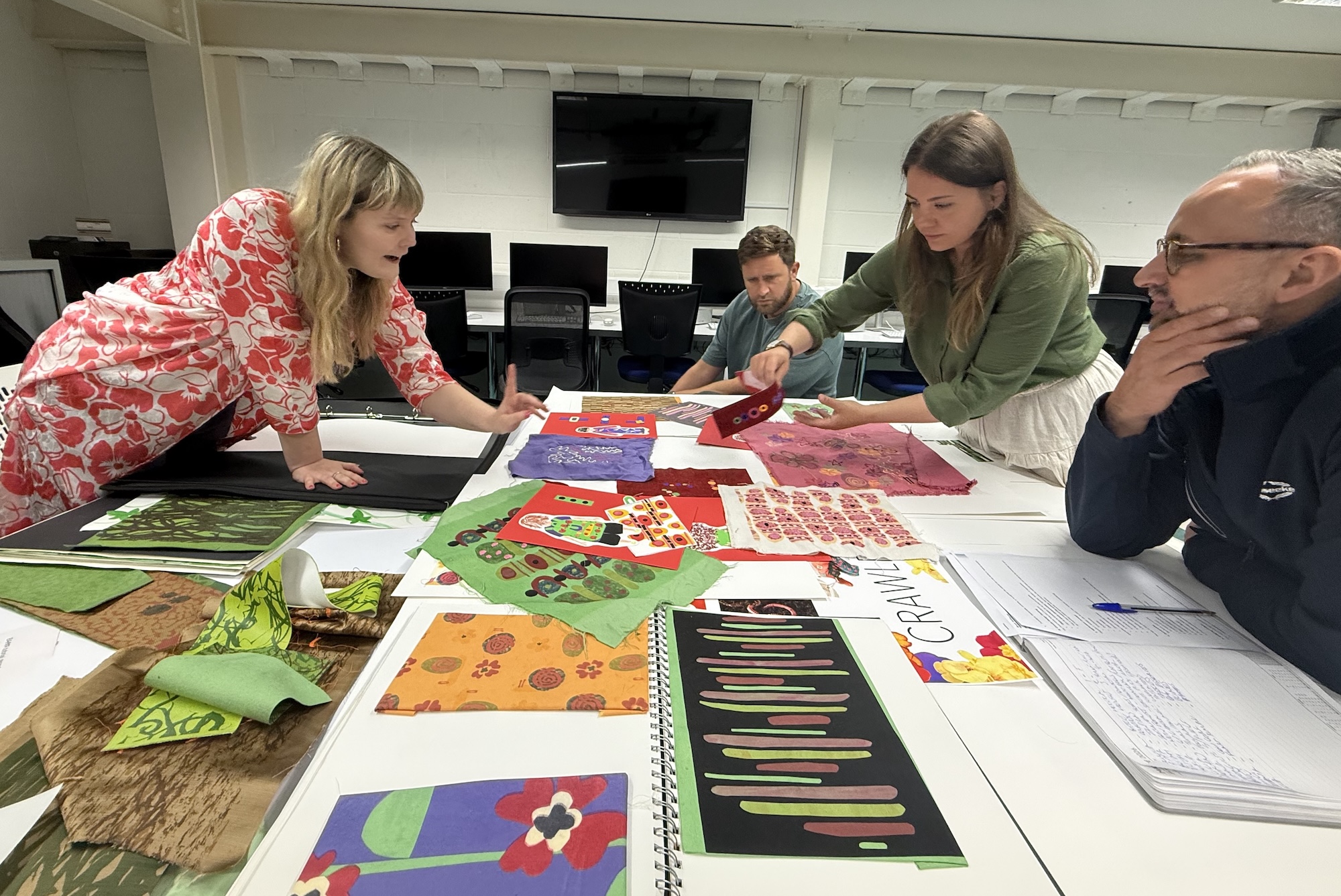 Two BA (Hons) Textiles Design students with two representatives from Sunseeker looking over various colourful textiles designs on a large white table.