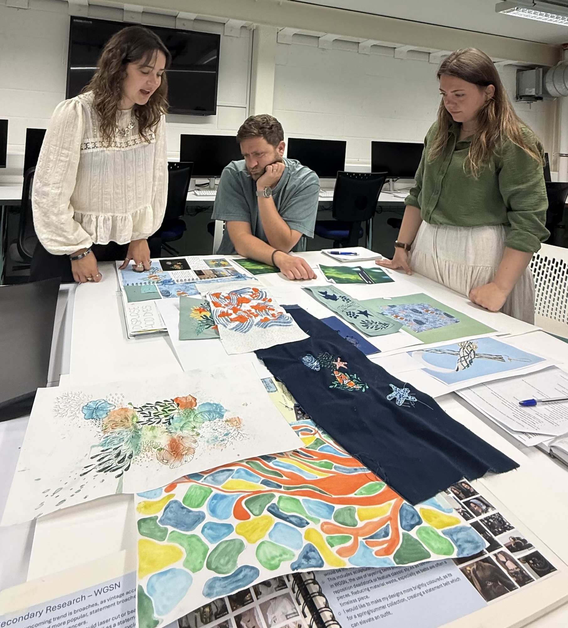 Three people reviewing various textiles designs around a table in a computer suite on Arts University Bournemouth campus.