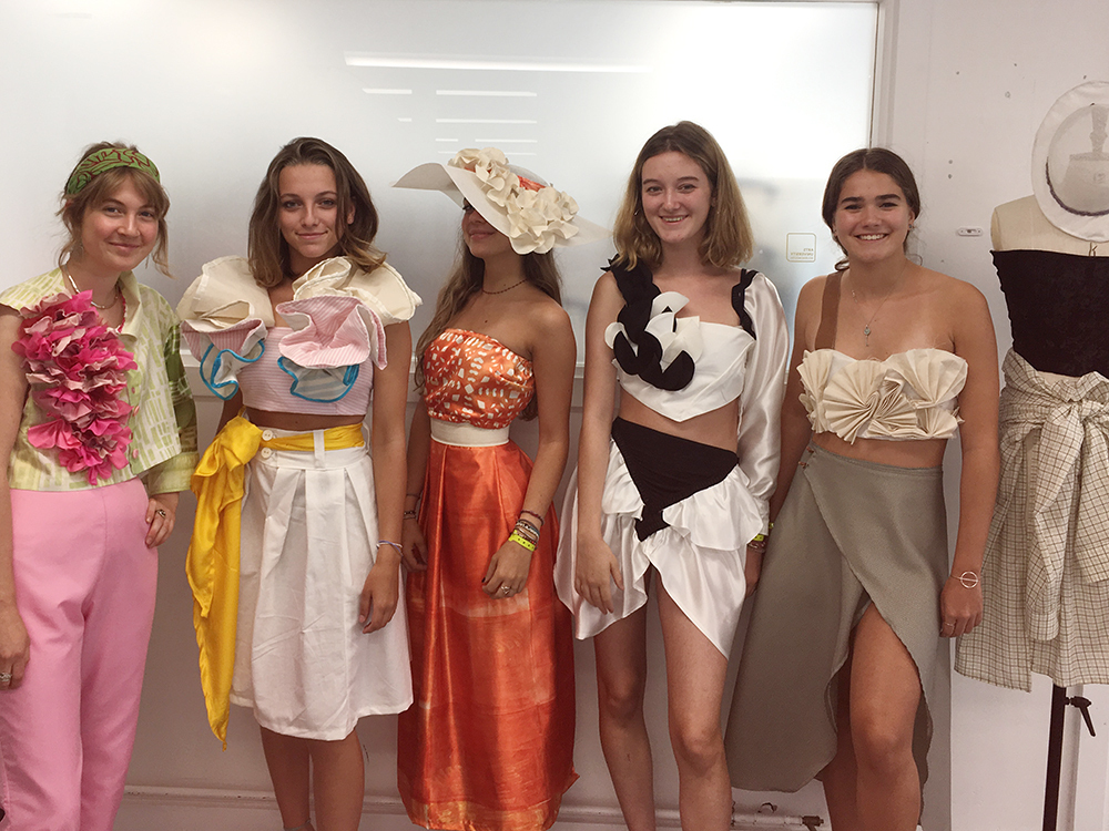 Five young women in elaborate costume dresses and hats posing together indoors, wearing colourful outfits with floral and fabric details.