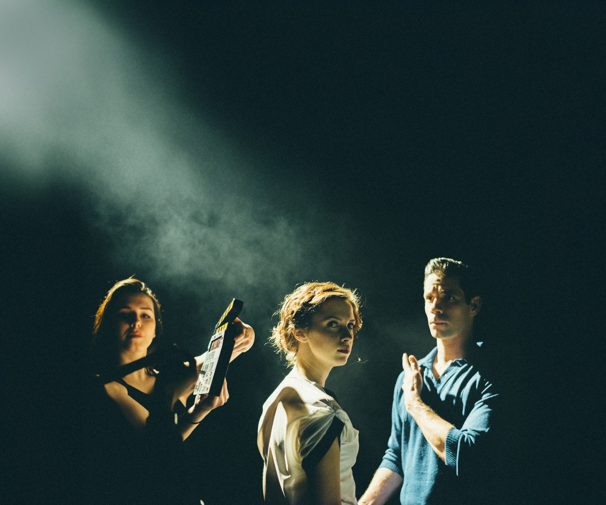 Three people standing in dramatic green-tinted lighting against dark background. Person on left holds papers, centre person wears glasses, right person in light blue shirt.