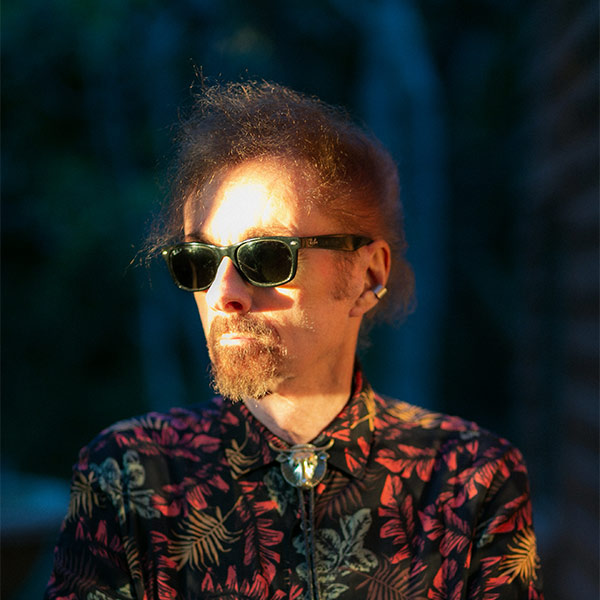 A man wearing dark sunglasses and a patterned shirt stands outdoors, with blurred trees in the background.