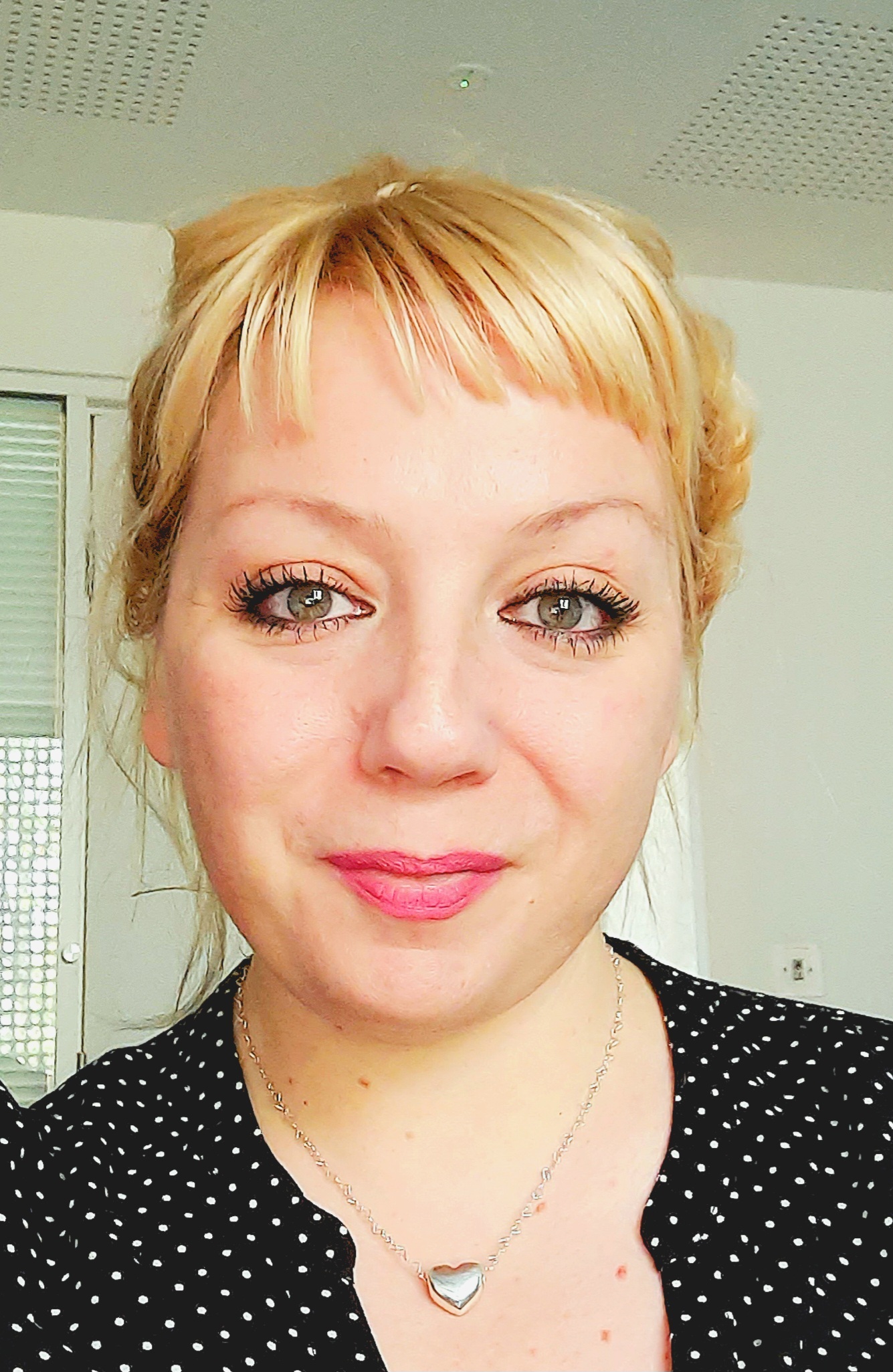 Woman with blonde hair and pink lipstick wearing black polka dot top and heart pendant necklace in bathroom with white tiles.