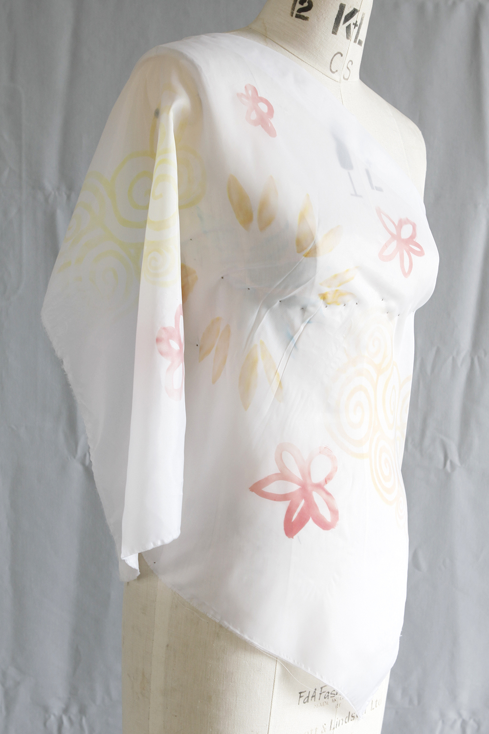 White sheer fabric with pink and yellow floral patterns draped on dress form mannequin against grey background.