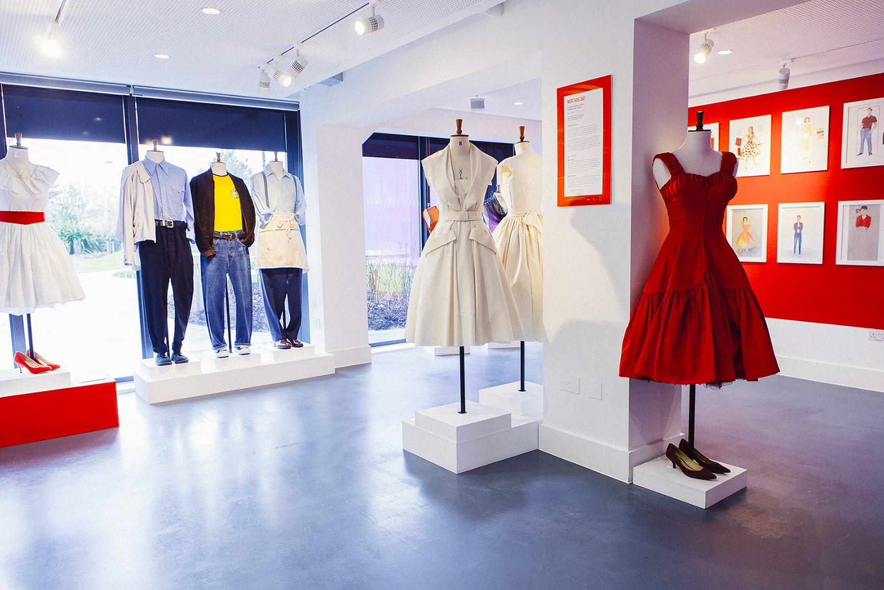 Three dresses on display on mannequins around a pillar in a gallery. Other outfits are on display further back.