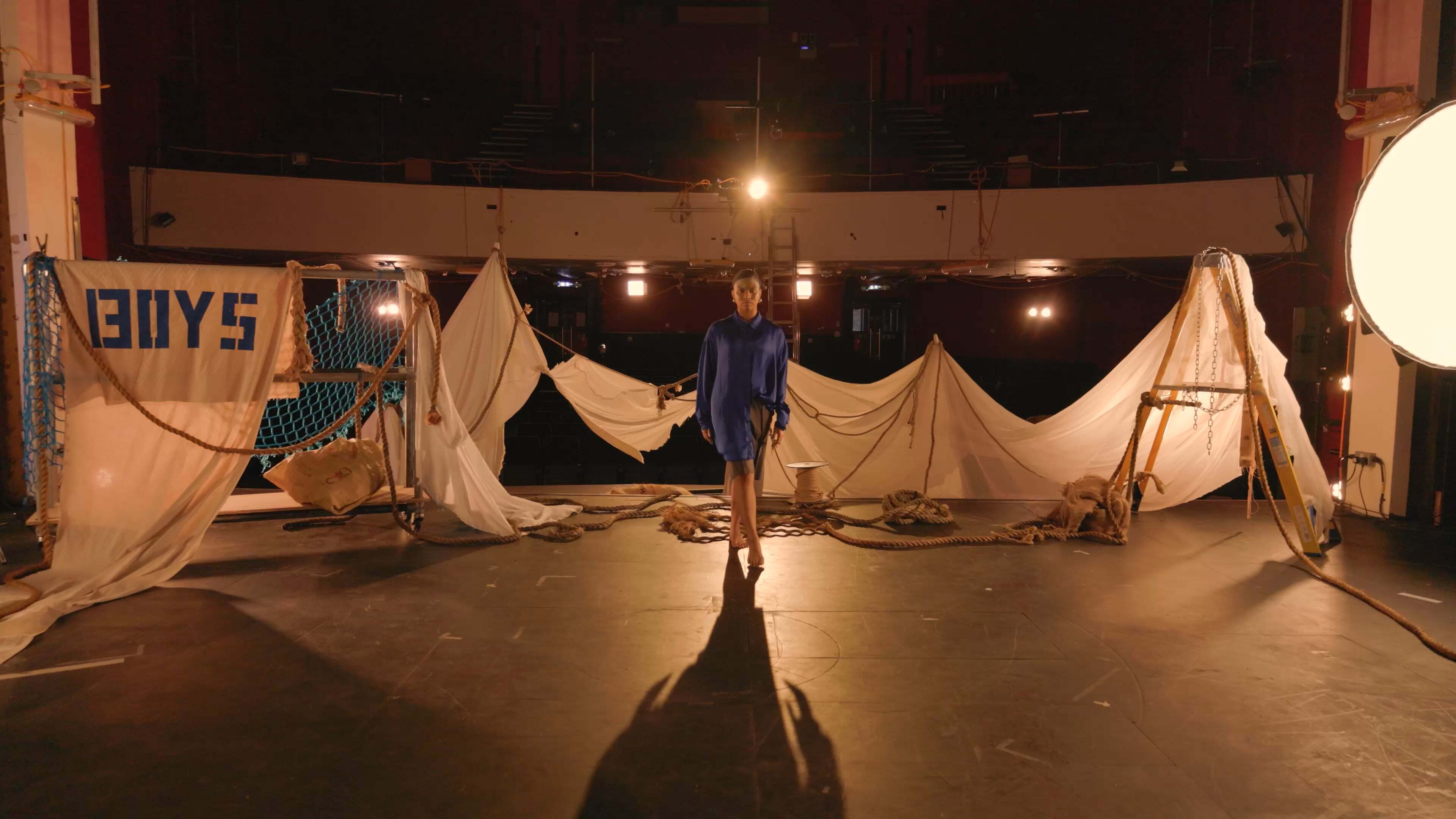 Person in blue dress standing on ship deck at night with white fabric draped between posts, warm lighting overhead.