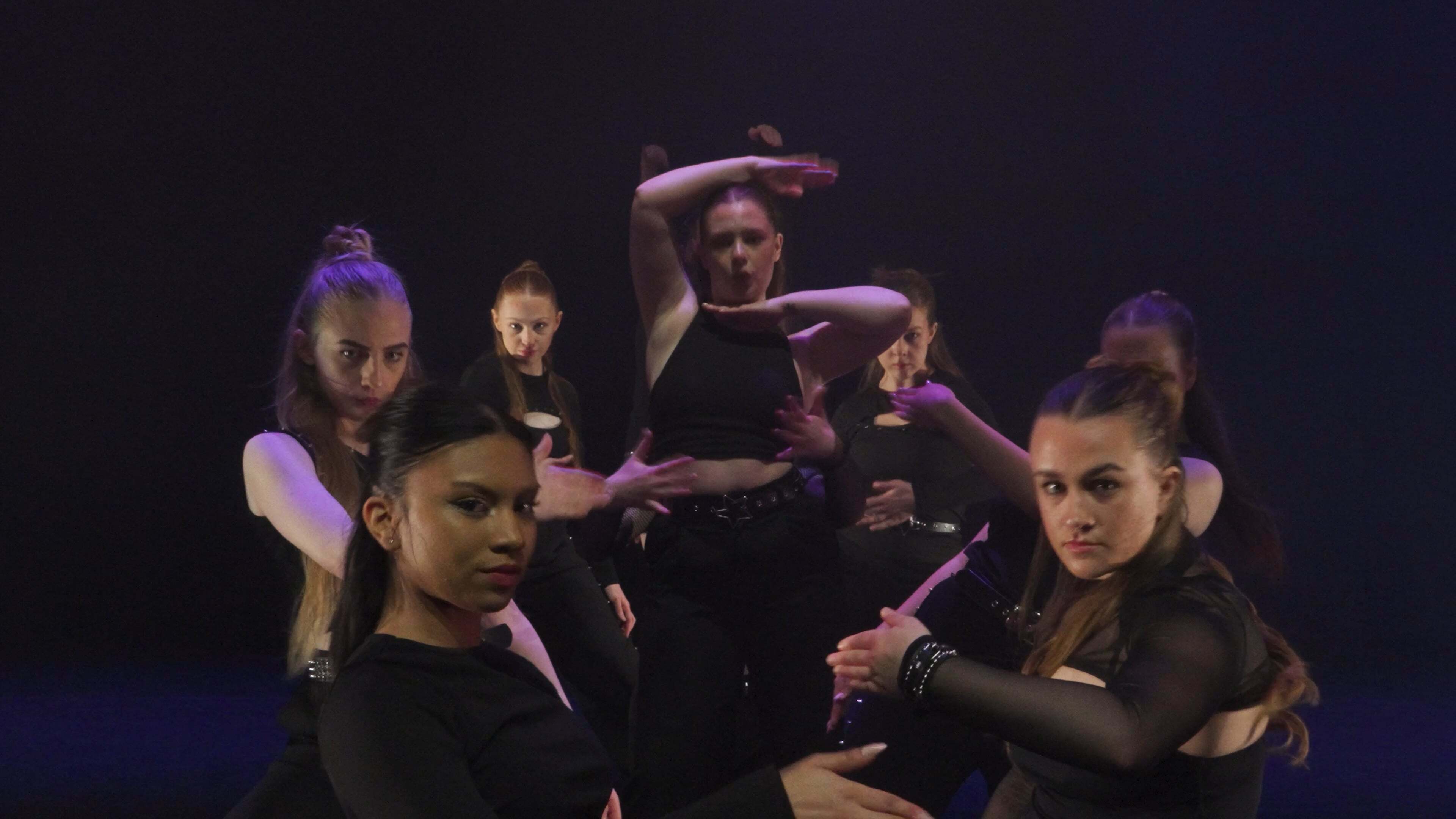 Group of dancers in black clothing performing on stage with purple and blue lighting against dark background.