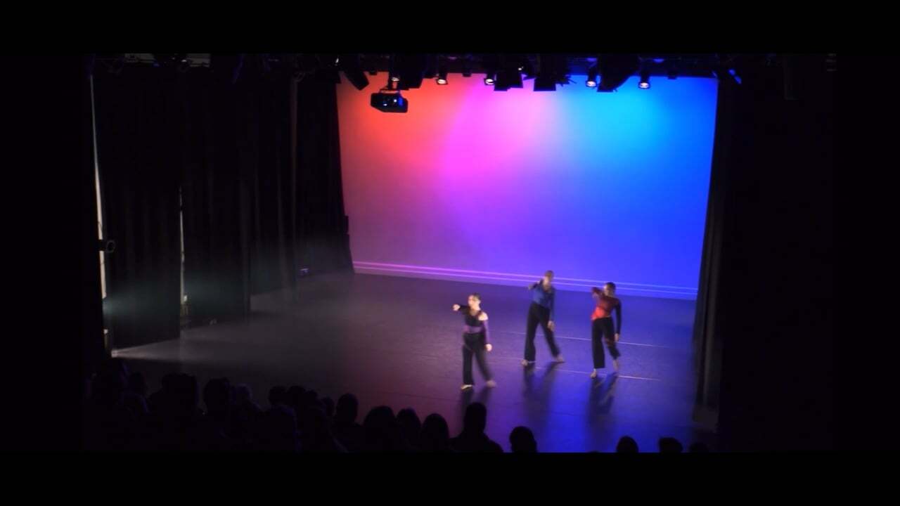 Three performers on stage with pink and blue gradient lighting backdrop, viewed from darkened auditorium with silhouetted audience.