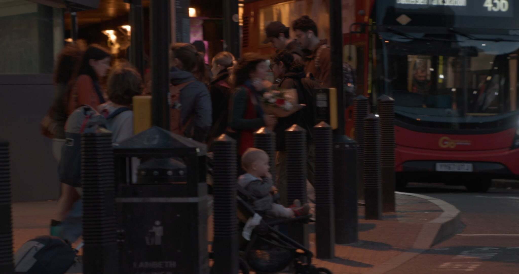 Evening street scene with people gathered near bus stop, red double-decker bus visible, warm lighting from shops and streetlights.
