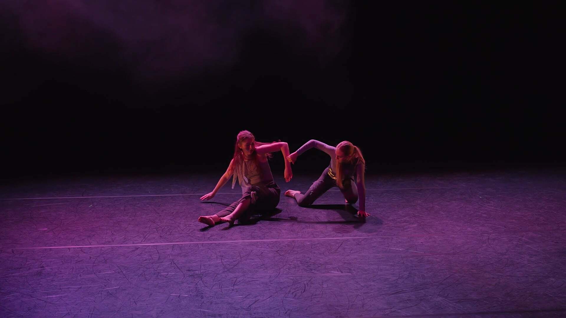 Two dancers on purple-lit stage floor against dark background, wearing light-coloured clothing, positioned close together in coordinated pose.