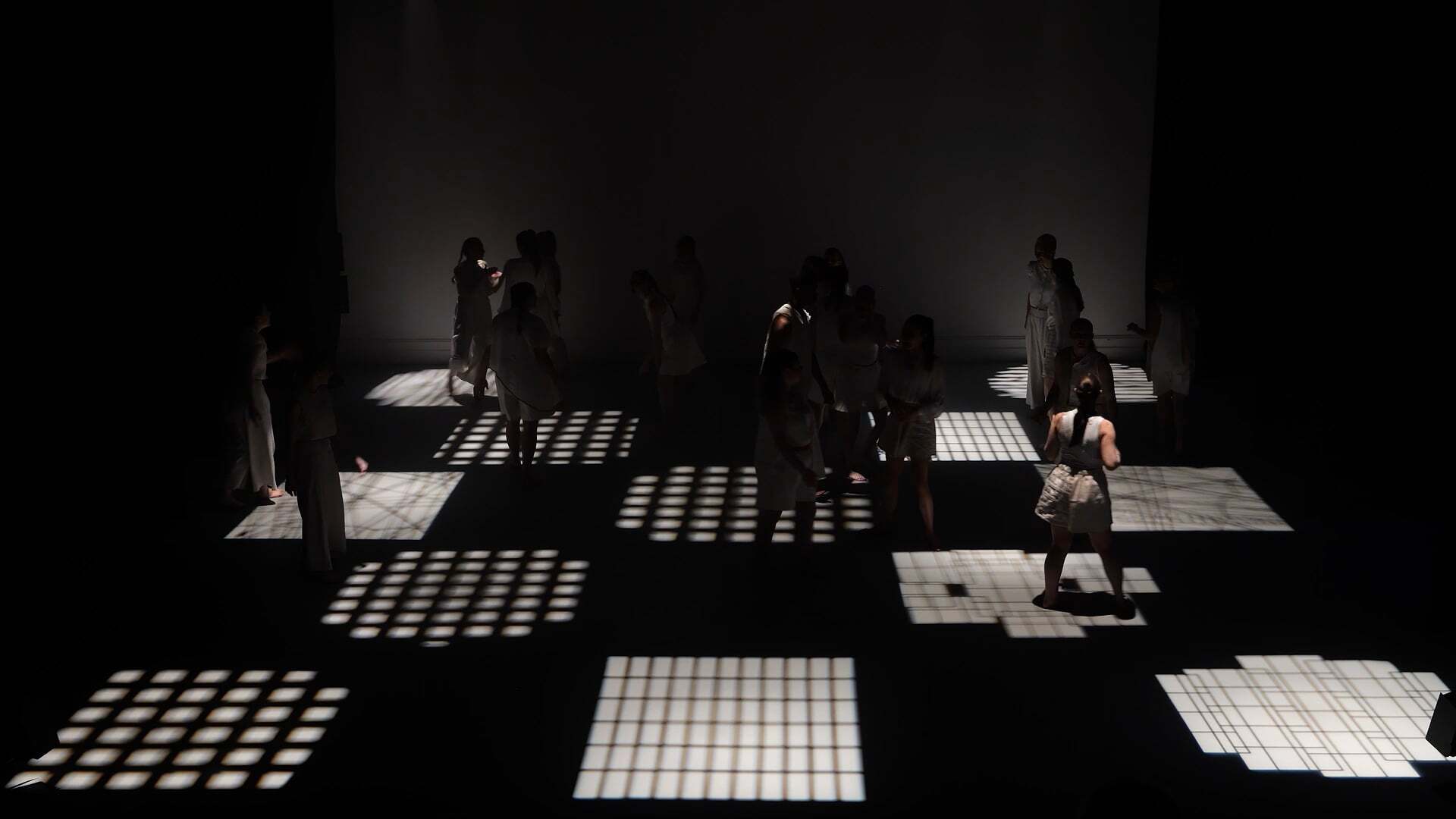 Dark room with illuminated white grid squares on floor, silhouettes of people standing and moving across the lit panels.
