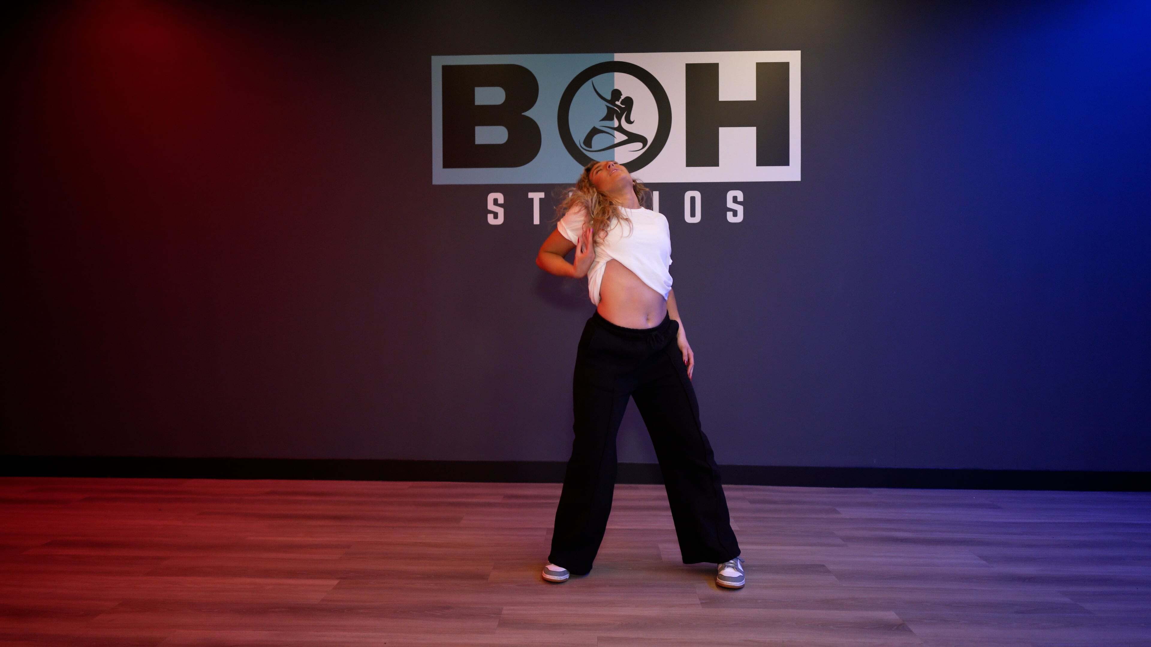 Woman in white crop top and black trousers dancing on wooden floor with red and blue lighting. "BOH STUDIOS" logo on wall behind.