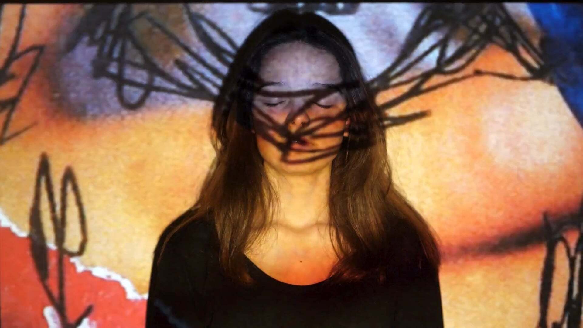 Woman with long brown hair wearing black top with orange, yellow, and blue collage projected onto her..