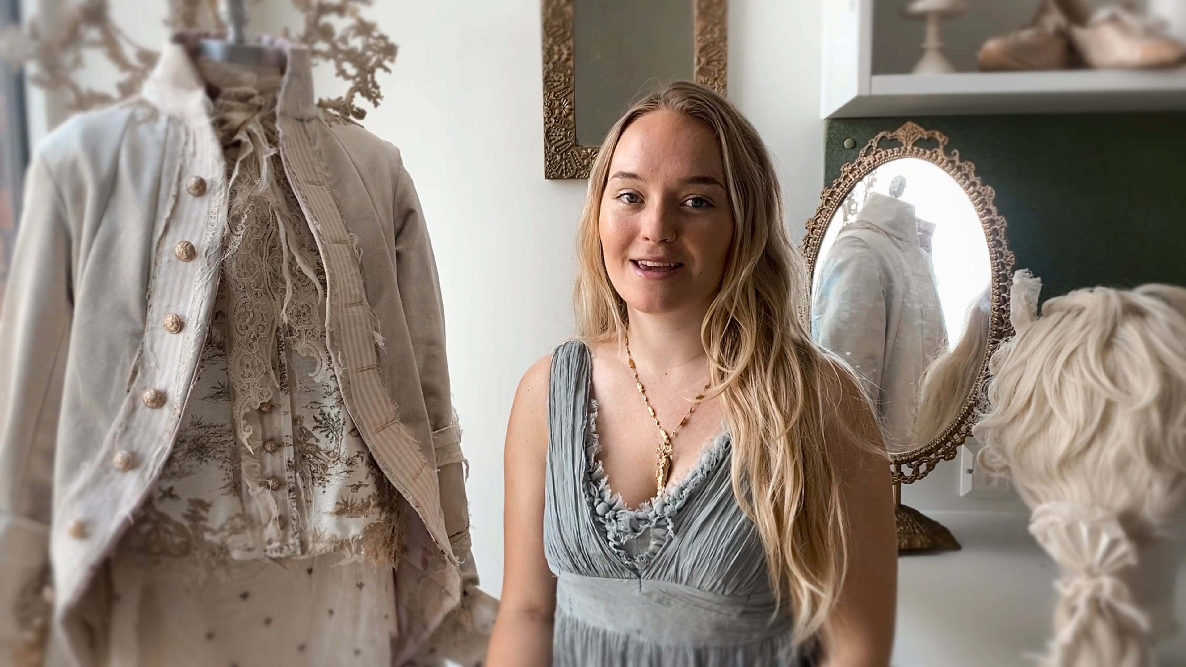 Young blonde woman in blue dress standing between white vintage clothing displays and ornate mirror in boutique interior.