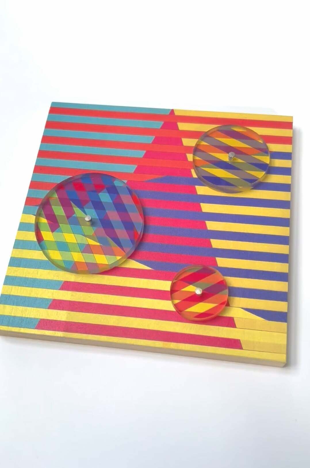 Colourful striped paper artwork with three transparent spheres creating optical effects. Red, yellow, blue, and green horizontal stripes.