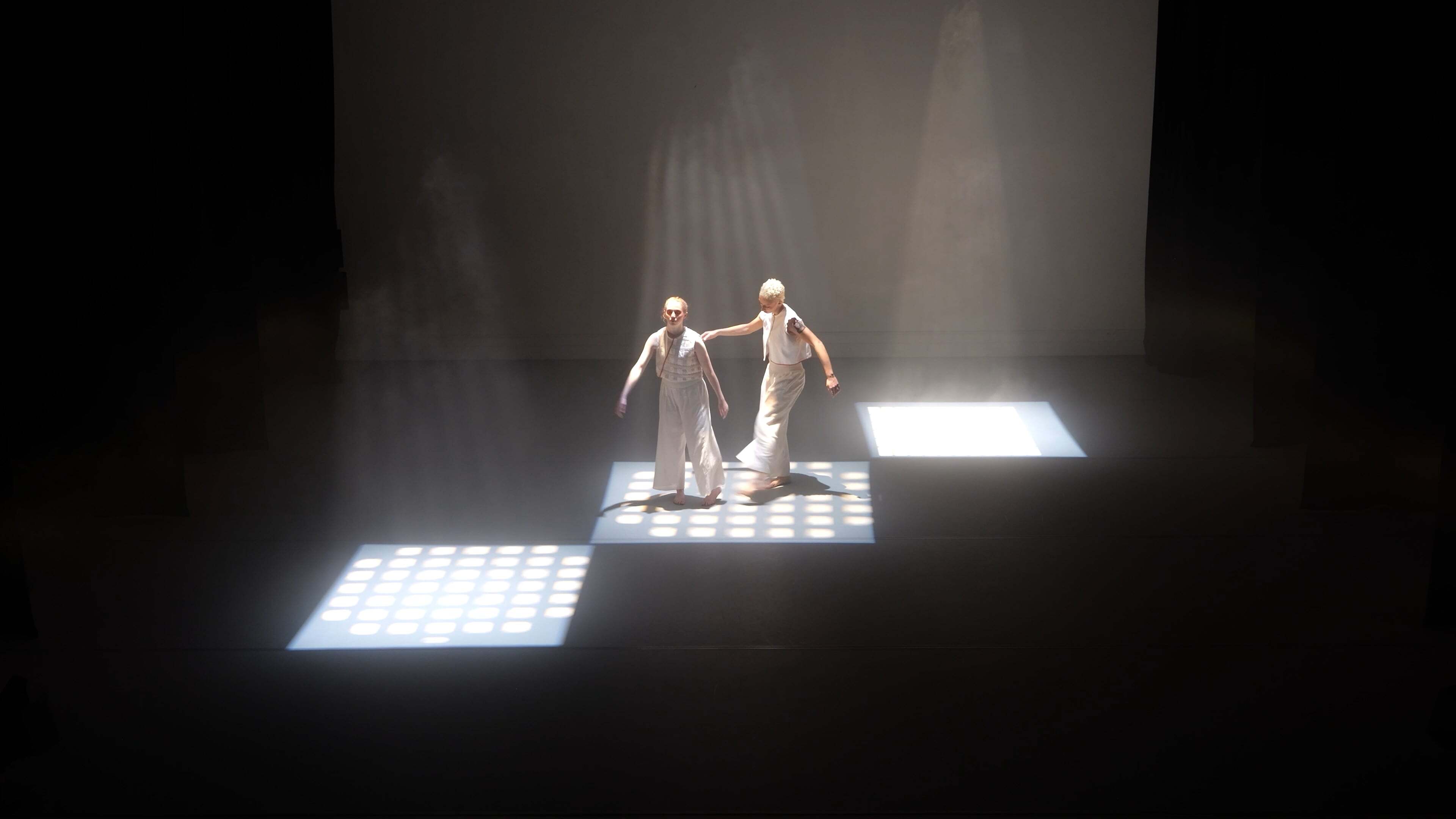 Two performers in white costumes on dark stage with dramatic overhead lighting creating bright rectangular pools of light.