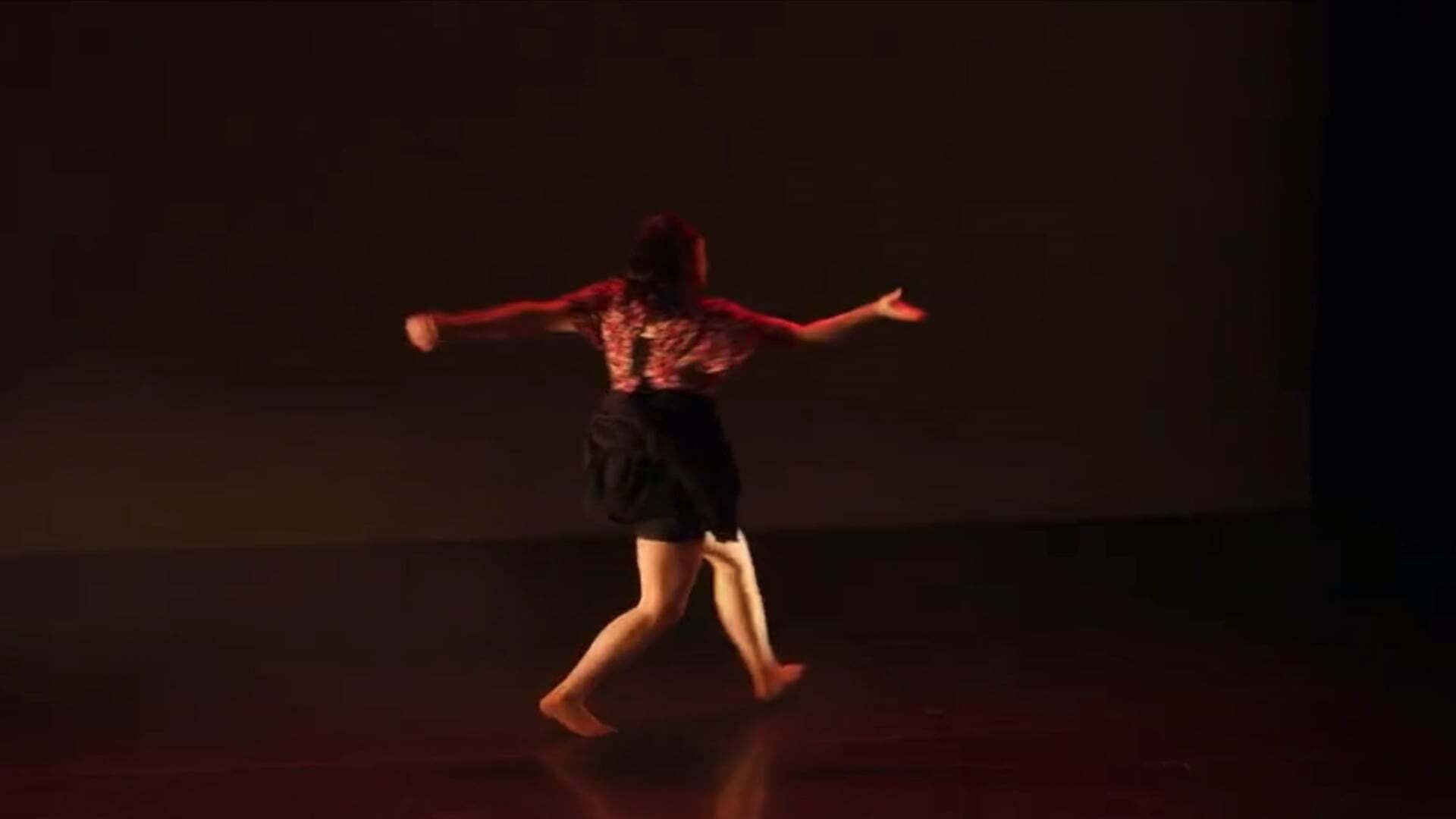 Barefoot dancer in red top and dark shorts with arms outstretched on dark stage, illuminated by warm stage lighting.
