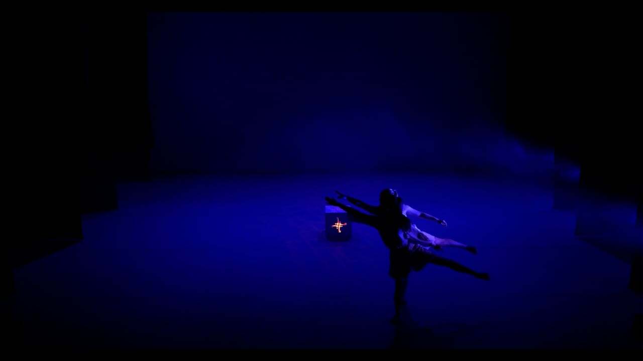 Dark silhouette of person in dynamic pose against bright blue illuminated stage, arms and legs extended outward.