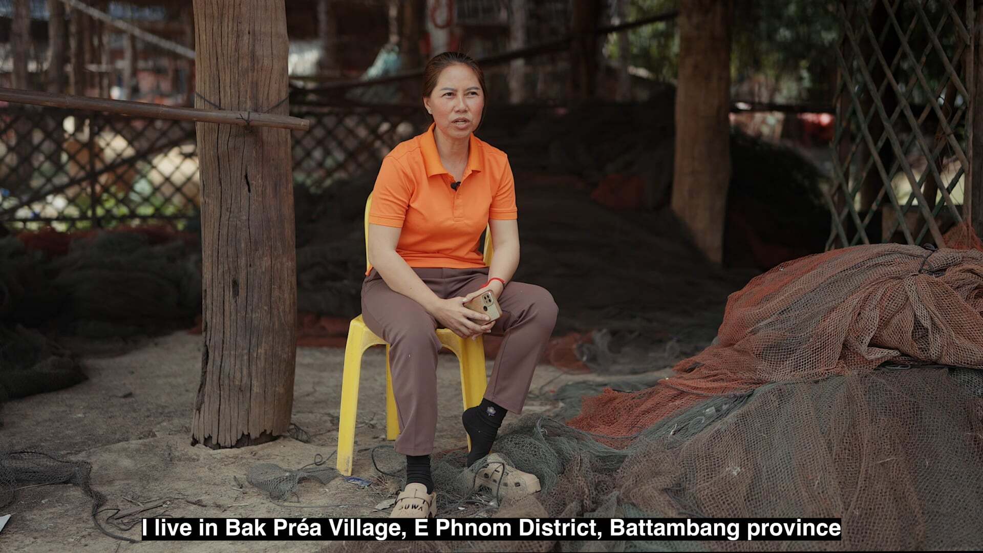 Woman in orange top sitting on yellow stool outdoors with wooden structures and lattice fencing in background. Caption visible.