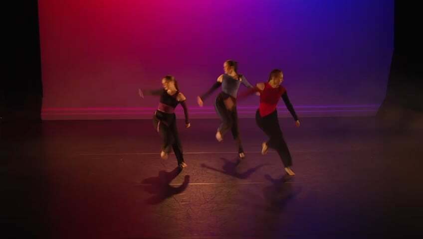 Three dancers performing on stage under red and purple lighting, wearing dark clothing with red accents.