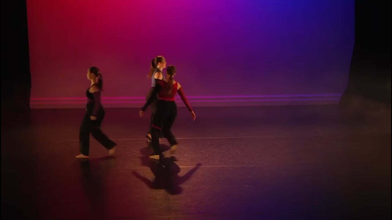 Two dancers on stage in dark clothing against purple and magenta gradient lighting backdrop.