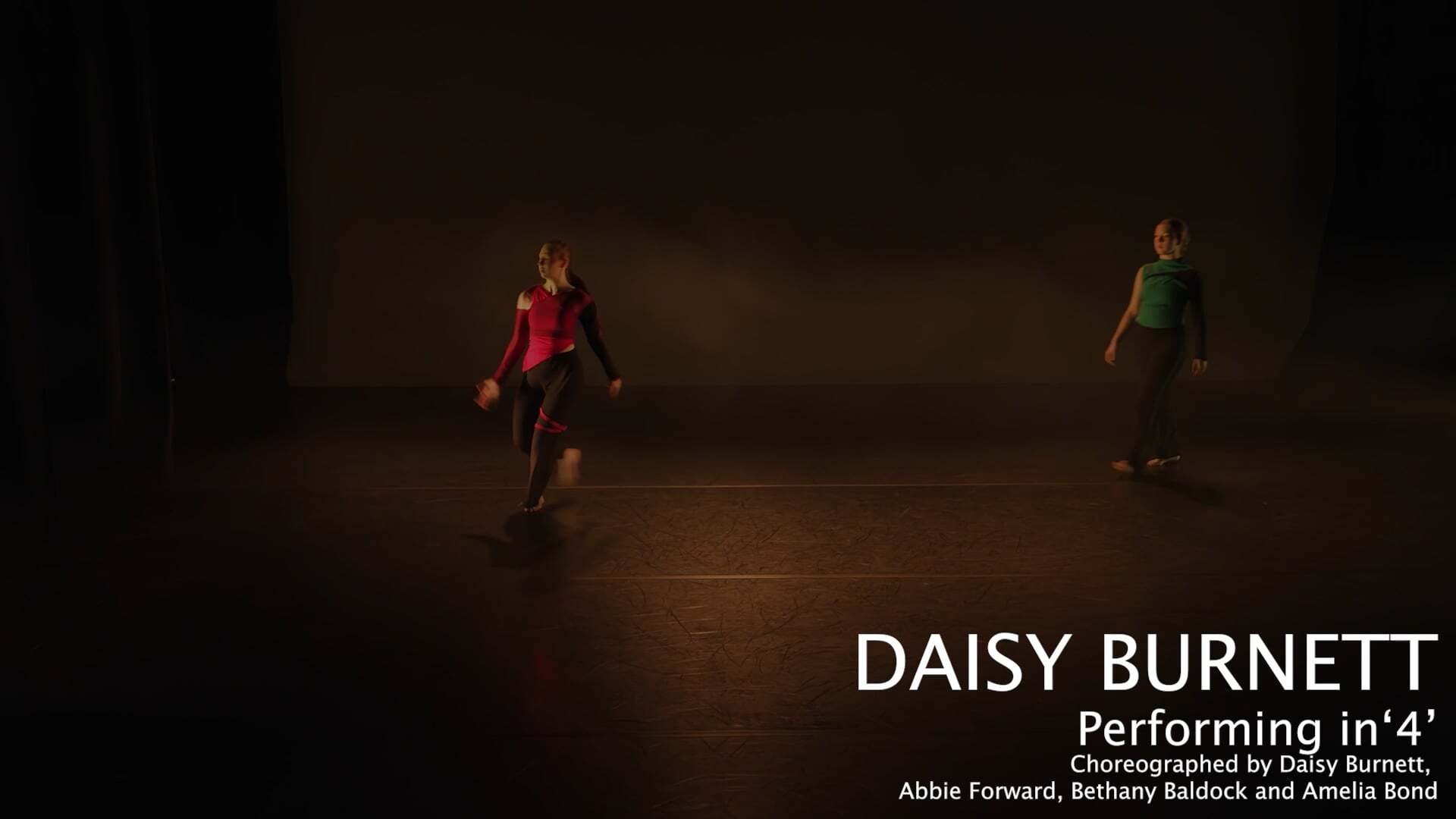 Two dancers in red and green clothing on dark wooden floor against brown wall background with white text overlay.