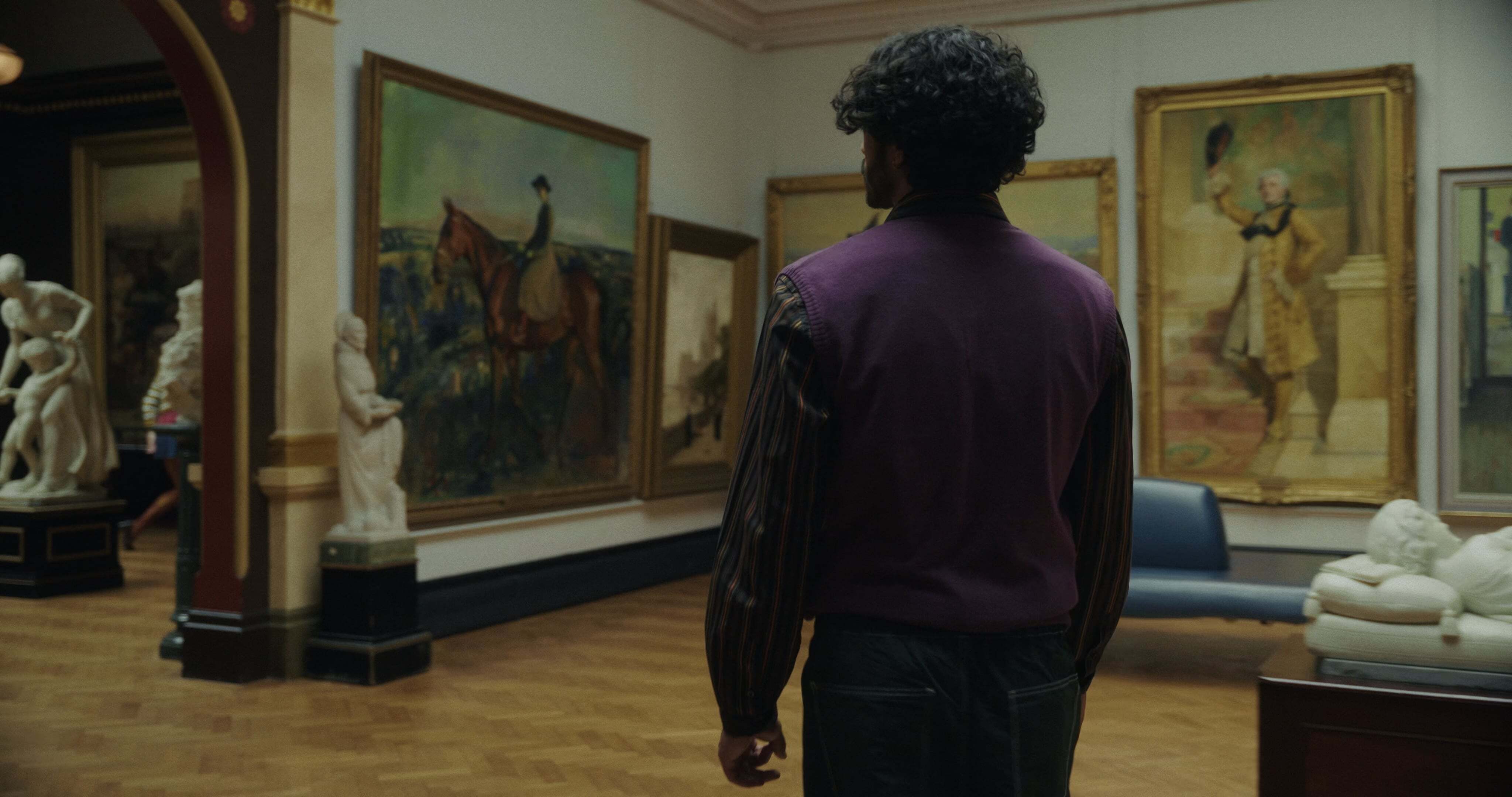 Person with curly hair in purple jumper standing in museum gallery with paintings and white sculptures on wooden floors.