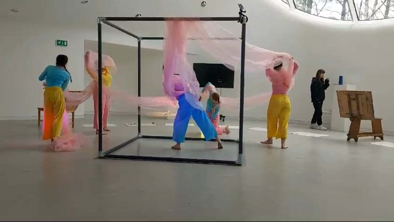 Performance art in white gallery space with performers in bright yellow and blue clothing, pink smoke, and black metal frame structure.