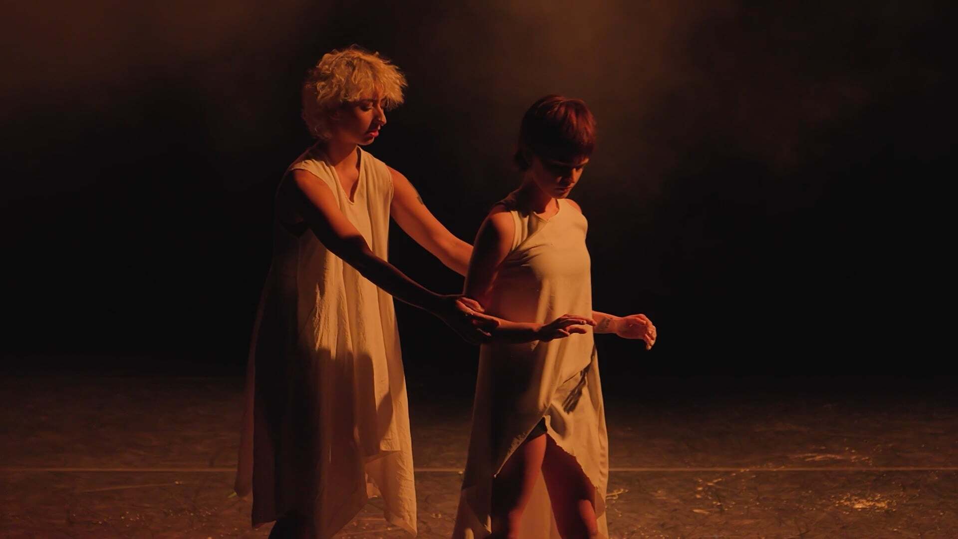 Two performers in white robes on wooden stage, one blonde figure restraining a dark-haired figure with rope, warm orange lighting.