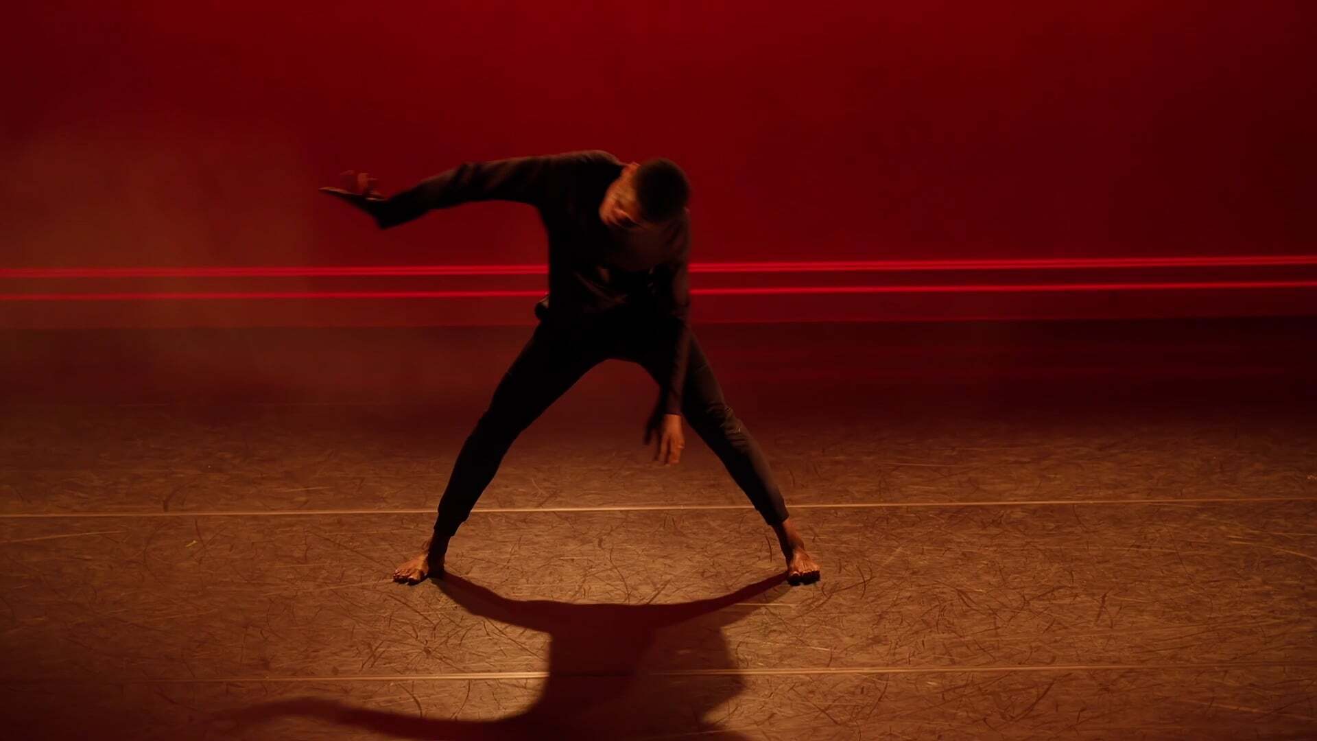 Dancer in black outfit performing dynamic movement in wide stance on wooden floor against red wall with horizontal lines.