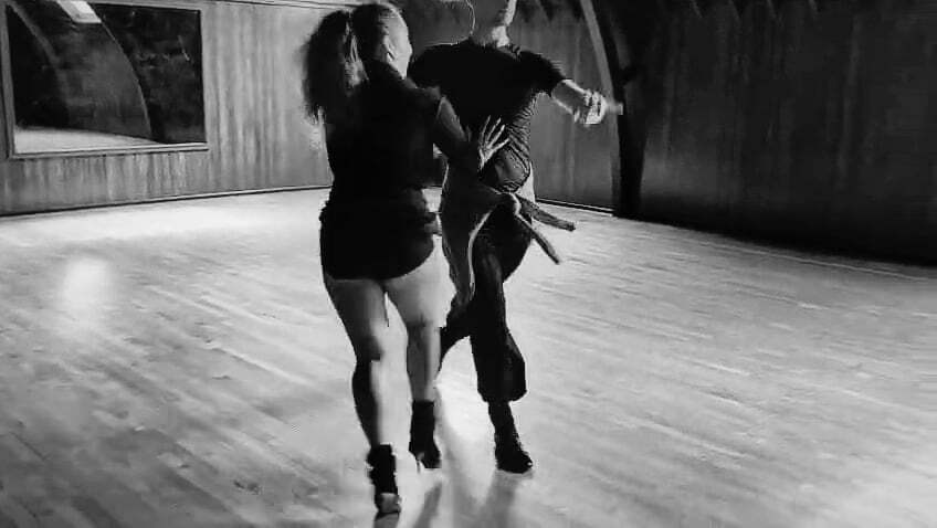 Two people dancing together in black and white, one in dark top and shorts, other in striped top and trousers, on wooden floor.